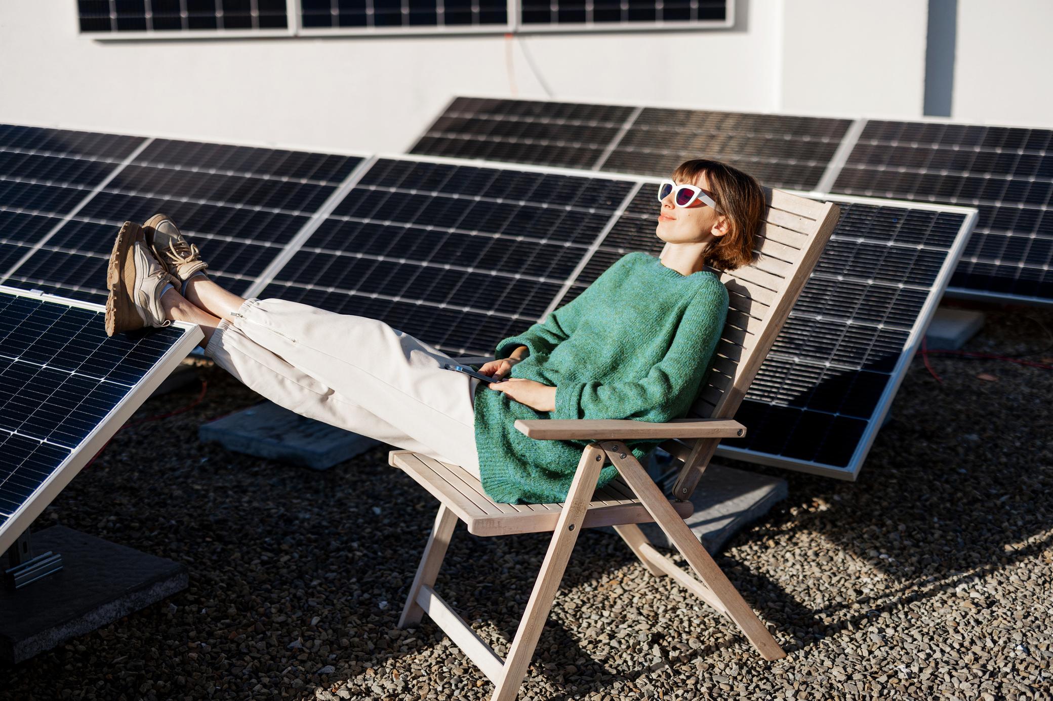 Junge Frau sitzt entspannt auf einem Liegestuhl und ruht sich auf einem Flachdach mit Solaranlage aus.