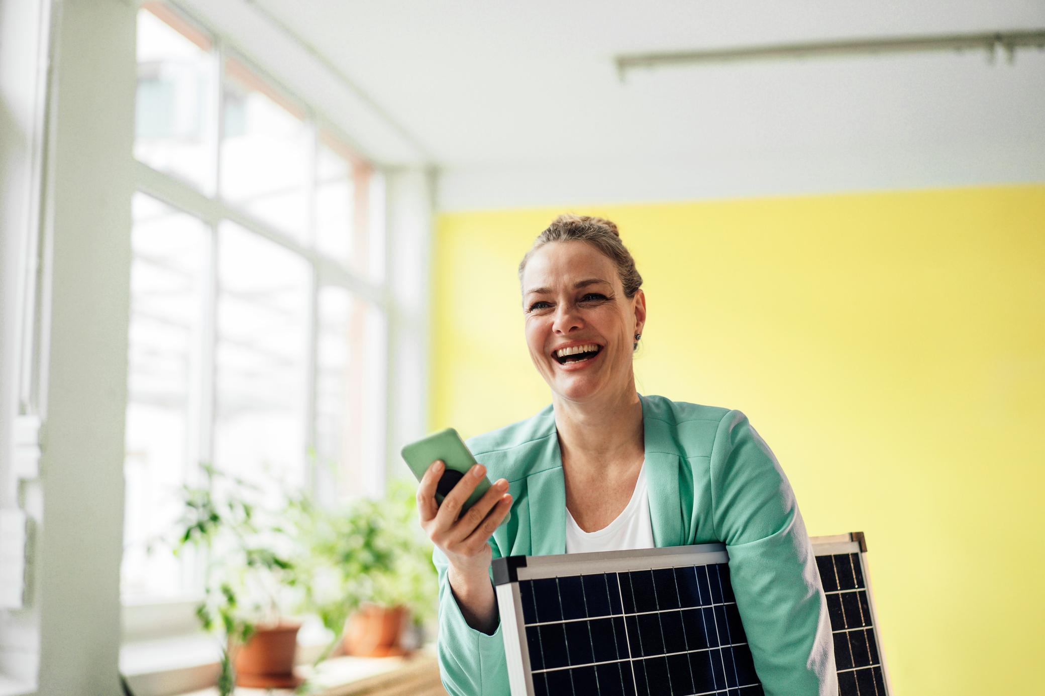 Eine lachende Frau die ein Handy in der Hand hält und ein kleines Solarmodul hält