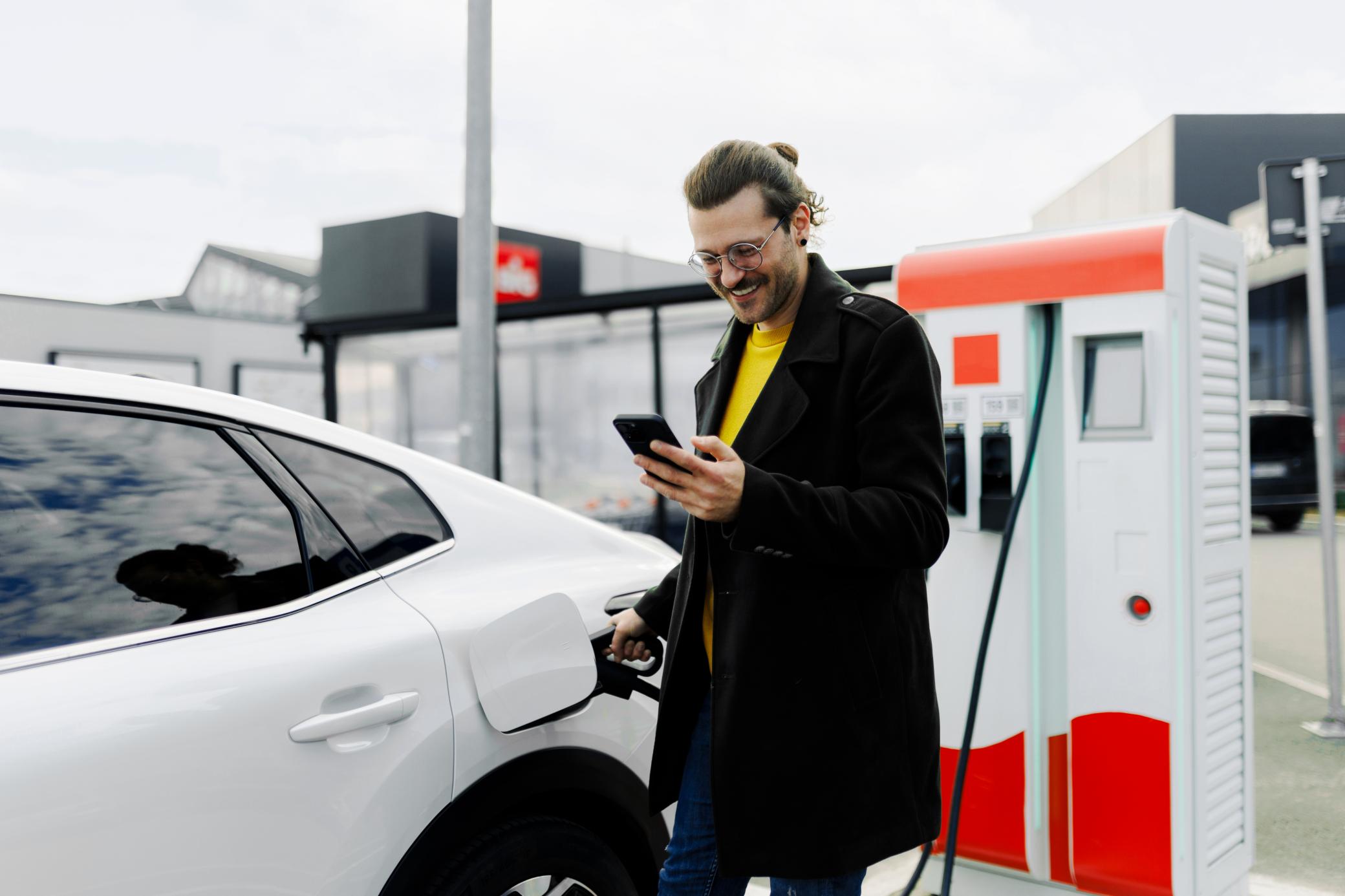 Ein lächelnder, junger Mann mit Smartphone lädt ein E-Auto an der öffentlicher Ladesäule