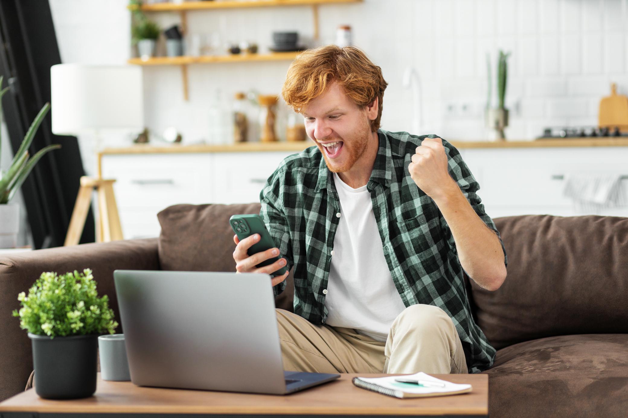 Jubelnder Mann im Homeoffice mit Laptop und Handy