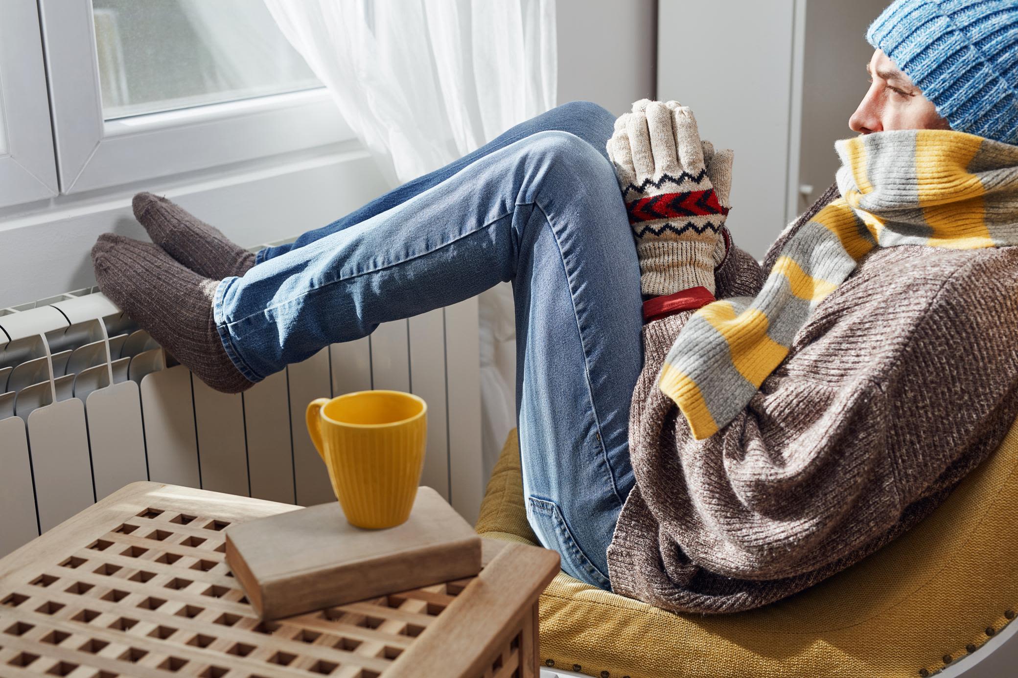 Mit Wärmestrom kannst du günstig und effizient zuhause heizen Mann sitzt mit Handschuhen, Mütze, Schal und Füßen auf der Heizung vorm Fenster, neben ihm steht eine Tasse Tee auf dem Tisch.