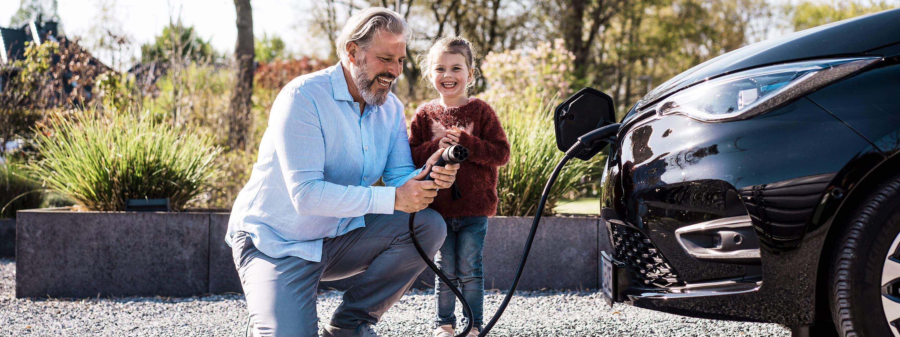 Mann mit Tochter, der an einem sonnigen Tag vor einem E-Auto einen Ladekabel in der Hand hält