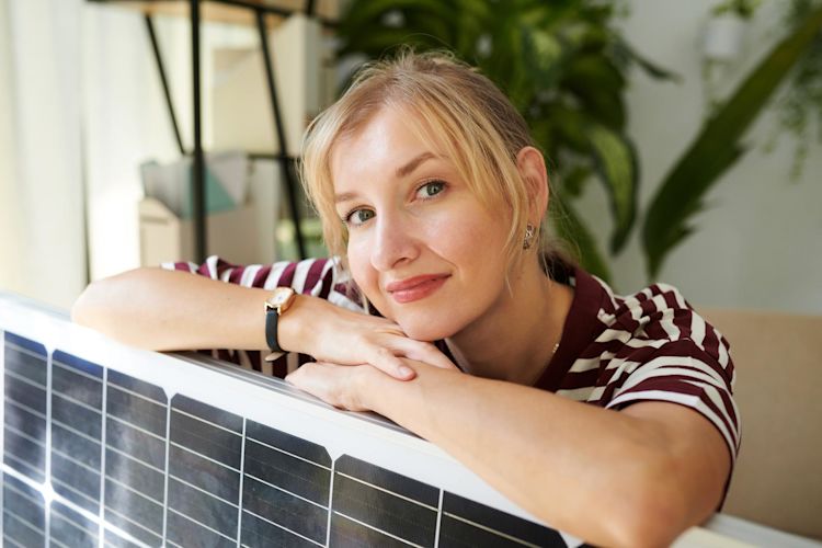 Strom mit Balkonsolaranlage kann so easy sein. Junge Frau mit blonden Haaren lehnt zufrieden über ihrem Solarpanel in ihrer Wohnung