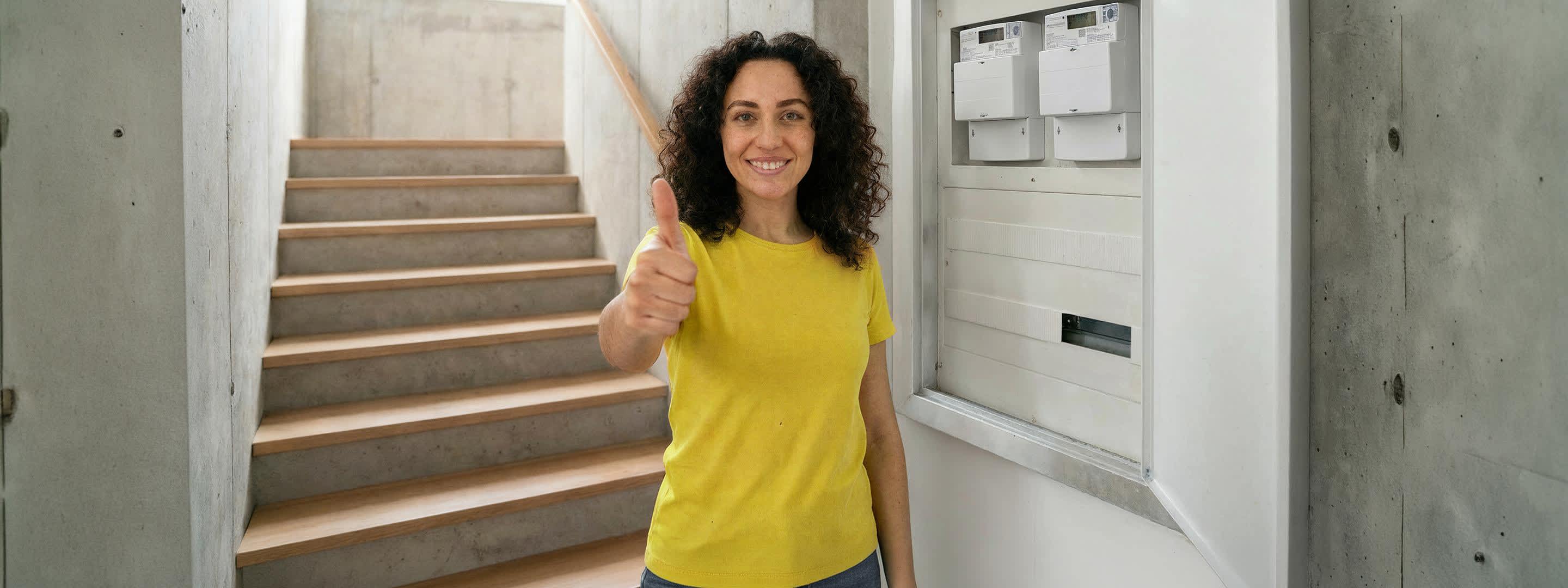 Eine Frau in gelbem Shirt steht neben Zählerkasten mit digitalem Stromzähler und zeigt lächelnd ihren Daumen hoch. Eine Frau in gelbem Shirt steht neben Zählerkasten mit digitalem Stromzähler und zeigt lächelnd ihren Daumen hoch.