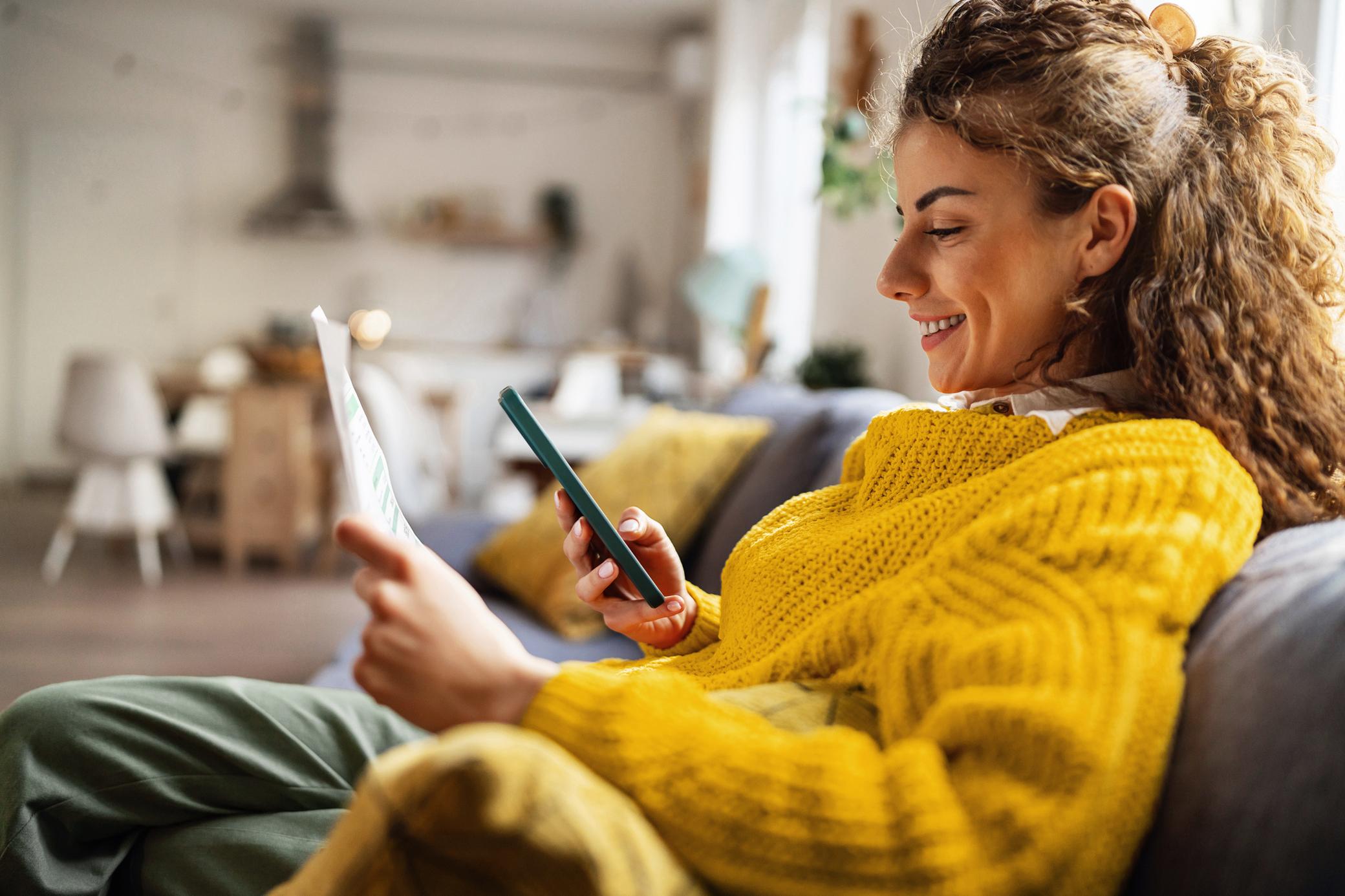Eine Frau sitzt auf dem Sofa und hält in der einen Hand einen Zettel, in der anderen ein Smartphone.
