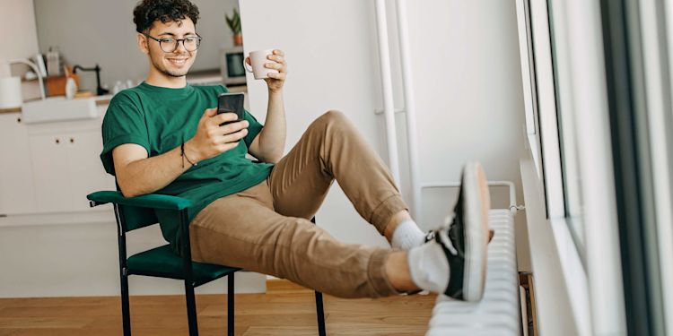 Mit der Wärmepumpen-Förderung zahlst du bis zu 70 % weniger Junger Mann sitzt lächelnd am Handy, mit den Füßen auf der Heizung