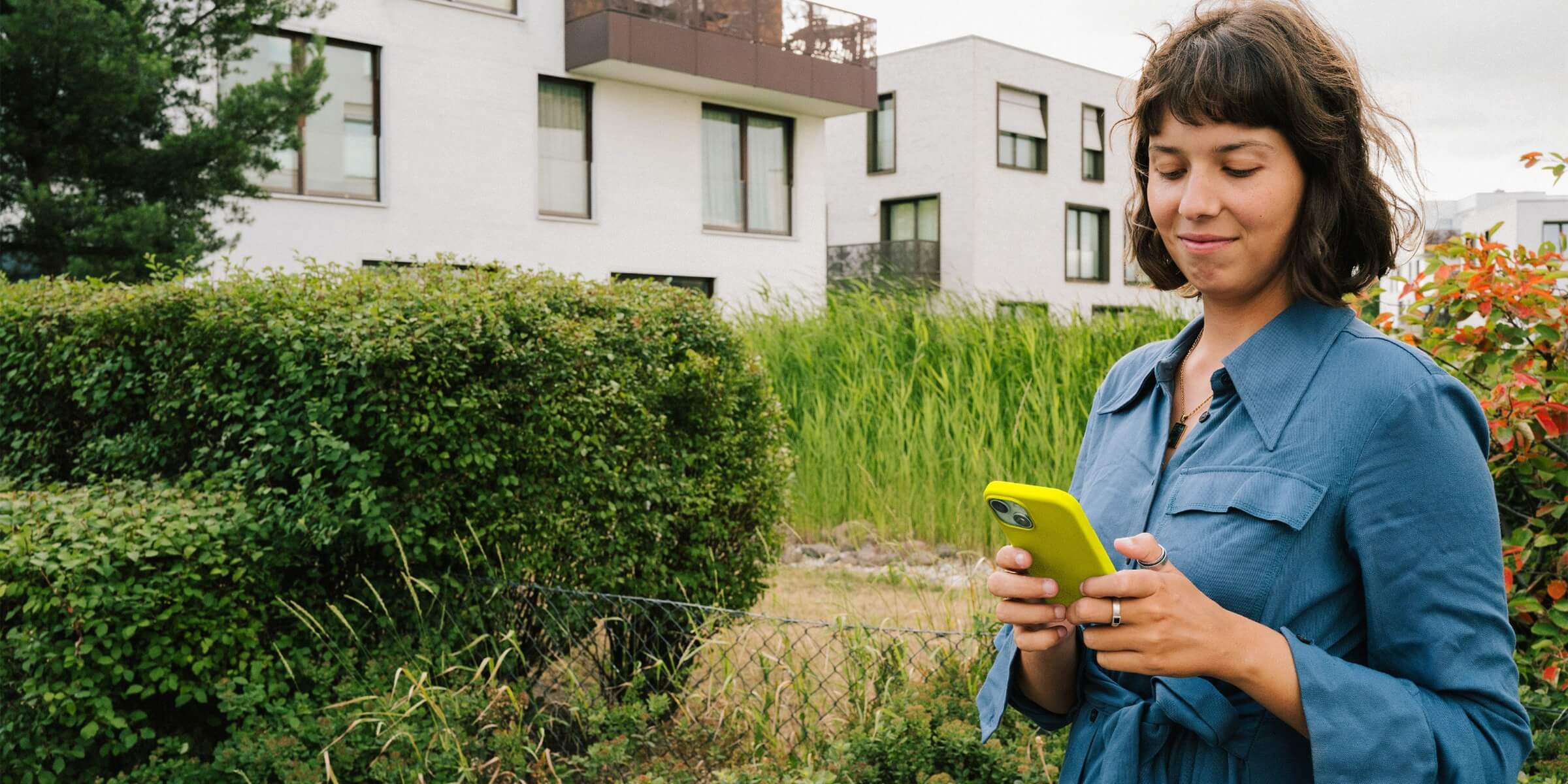 Auch im Mehrfamilienhaus kannst du erneuerbare Energien nutzen Junge Frau steht mit einem gelben Handy im Grünen vor einem Mehrfamilienhaus