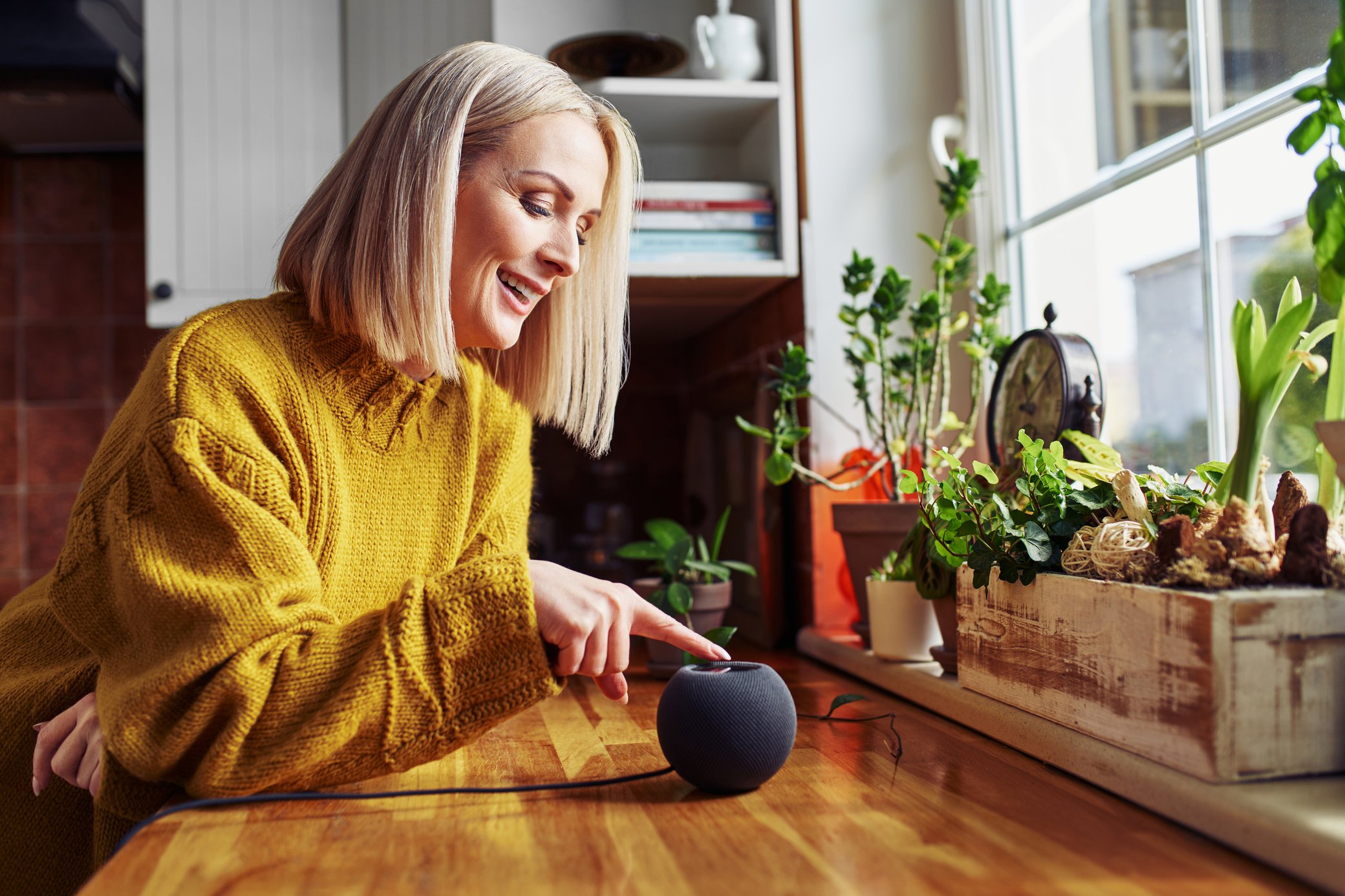 Frau in gelbem Pullover tippt auf einen Smart Speaker, in einer Küche mit Pflanzen vor dem Fenster