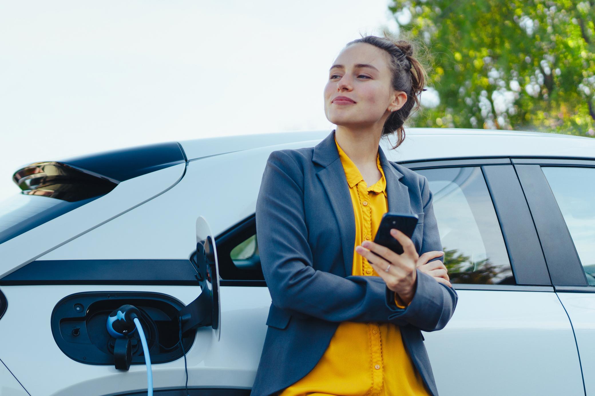 Junge Frau mit Smartphone beim Laden ihres Elektroautos