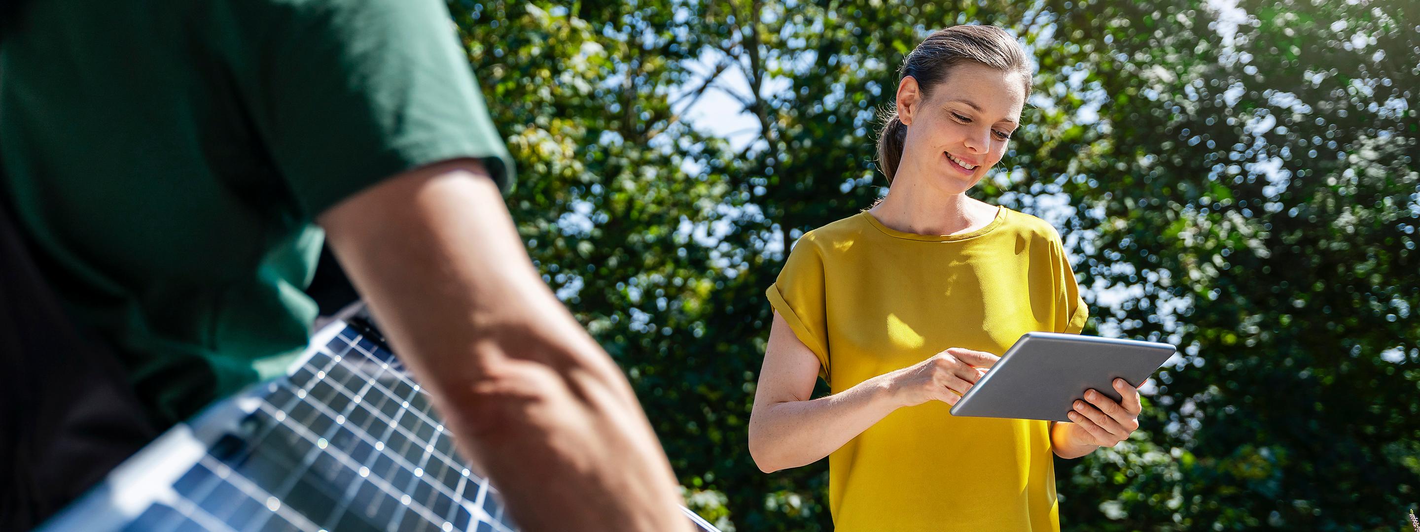 Frau im gelben Shirt steht in der Natur vor grünen Bäumen mit Tablet in der Hand und Mann mit grünem Shirt trägt ein Solarpanel