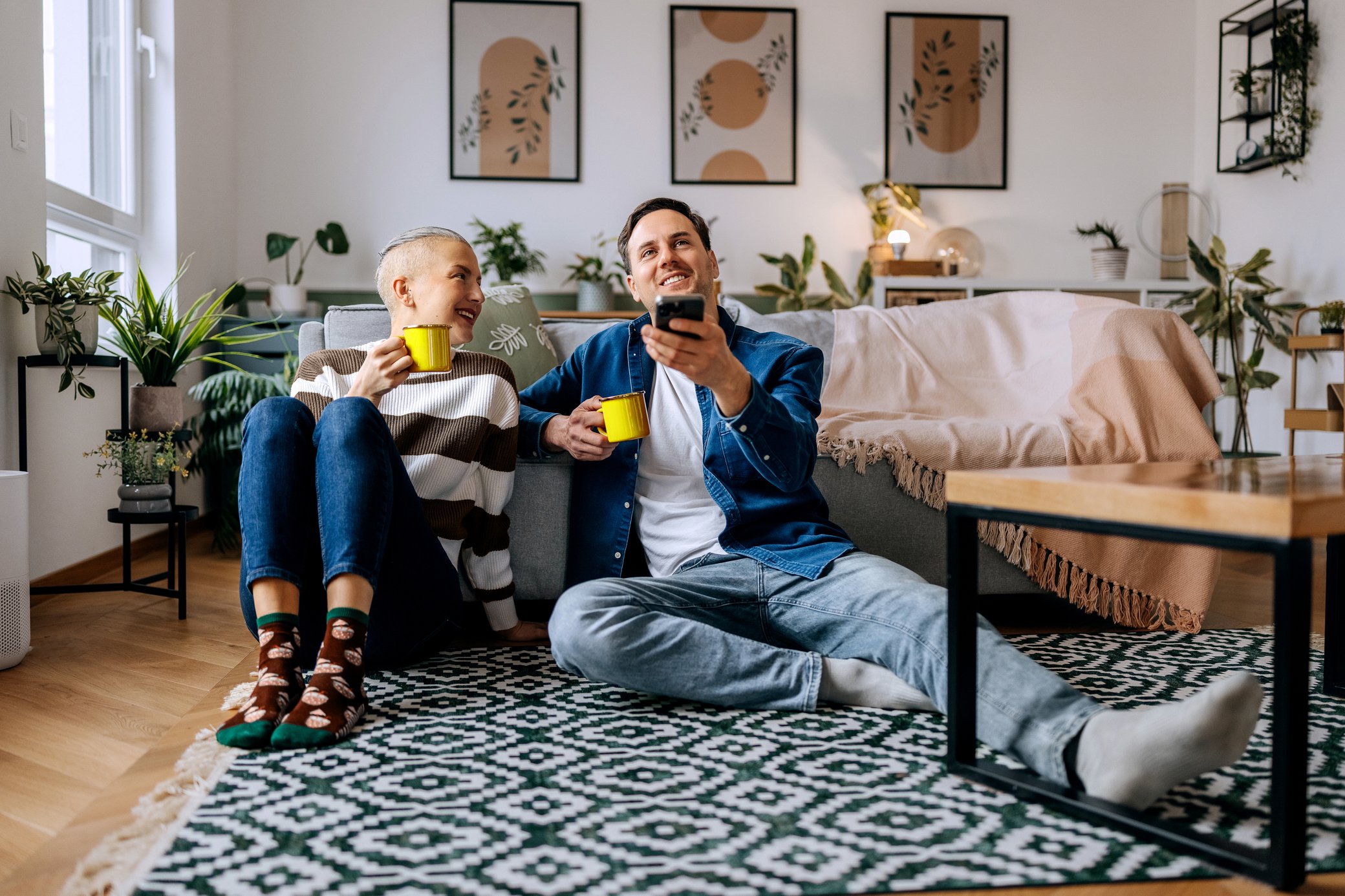 Ein junges Paar sitzt mit einer Tasse Tee in der Hand auf dem Boden und steuert ein Smart-Home-System mit dem Smartphone
