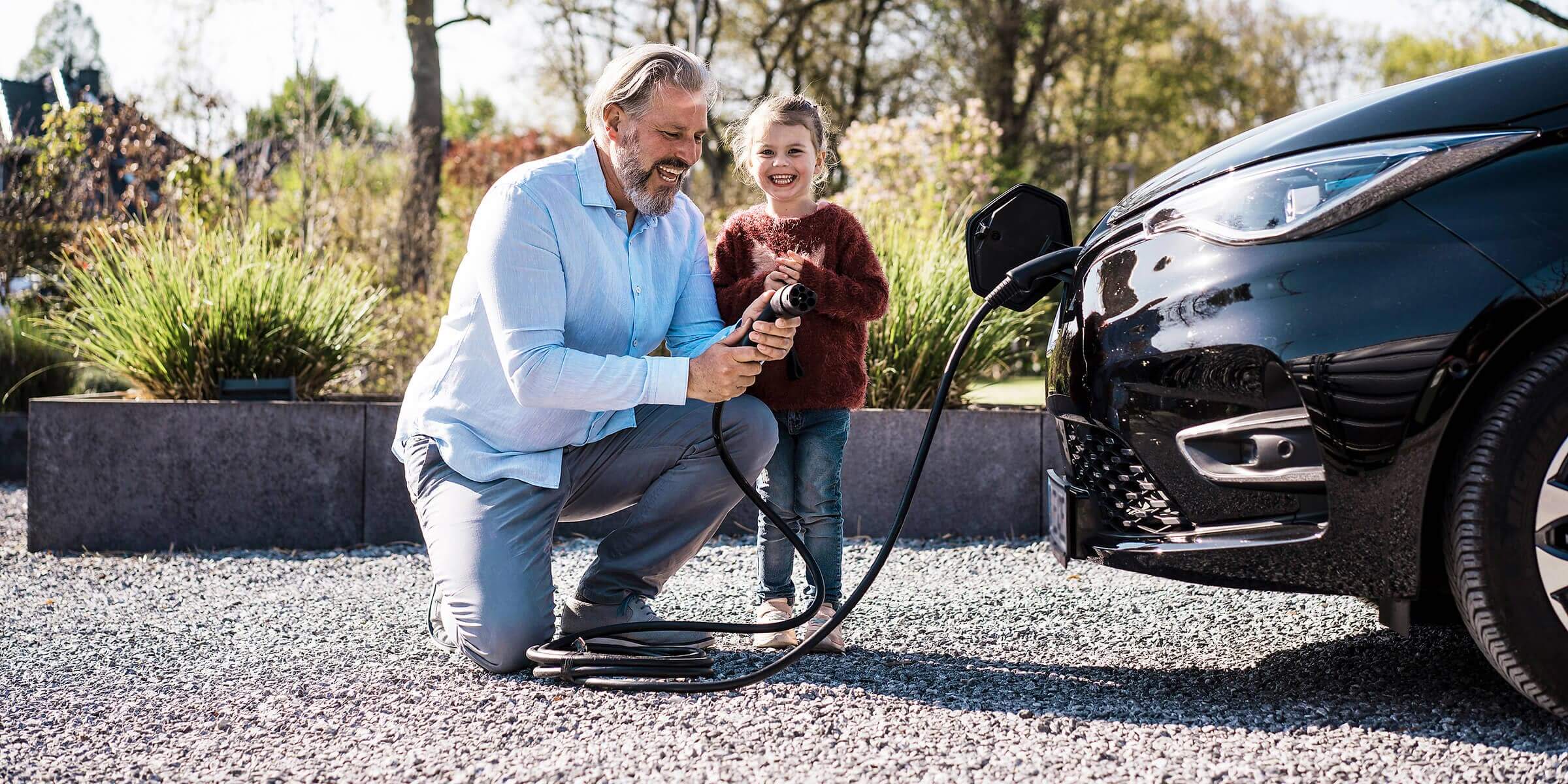 Mit Heim-Energie-Management-Systemen holst du mehr aus deinen Geräten raus Mann mit Tochter, der an einem sonnigen Tag vor einem E-Auto einen Ladekabel in der Hand hält