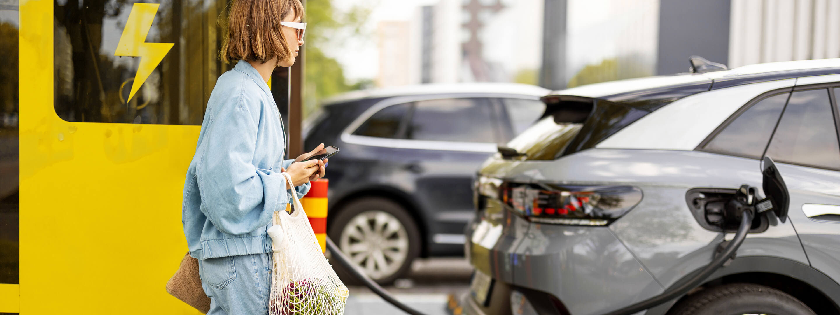 E-Auto Kosten im Überblick - Von Kauf bis Wartung Frau mit Handy und Lebensmitteln wartet auf Laden des Elektroautos