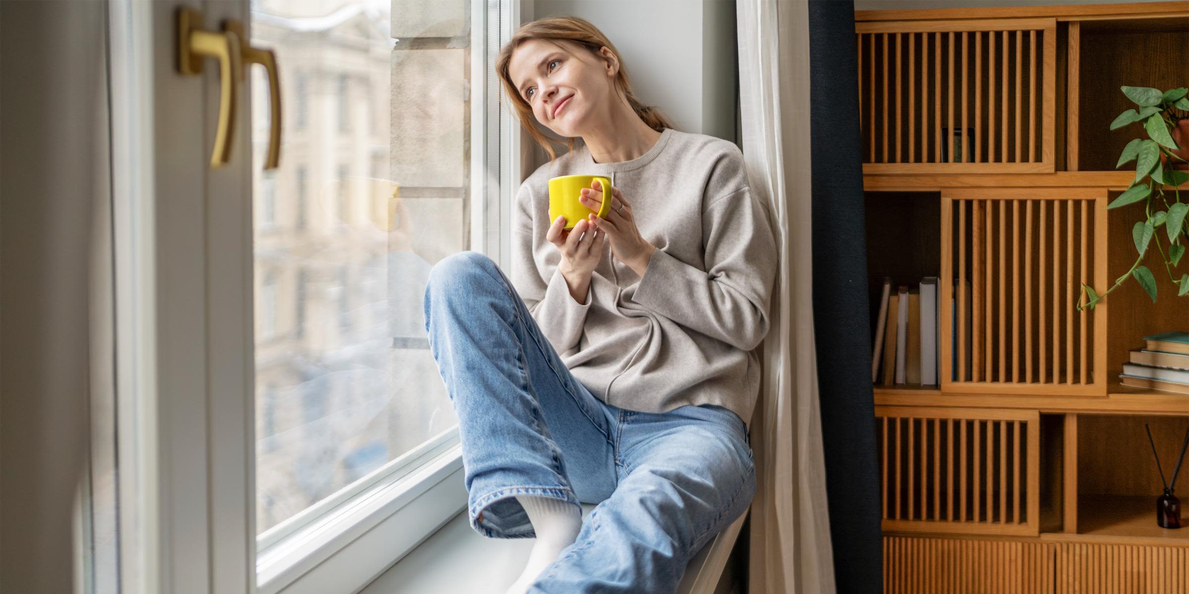Immer up-to-date beim Gaspreis? Wir haben alle Infos. Frau sitzt auf Fensterbank über Heizung und hält eine gelbe Tasse in der Hand