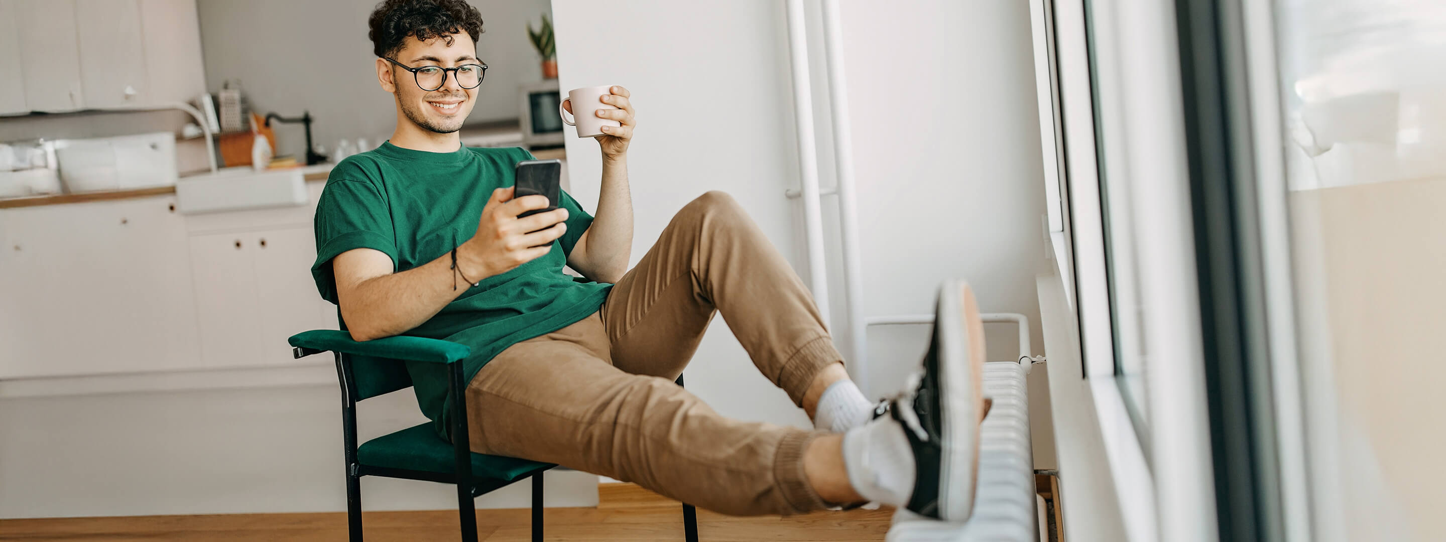 Mit der Wärmepumpen-Förderung zahlst du bis zu 70 % weniger Junger Mann sitzt lächelnd am Handy, mit den Füßen auf der Heizung
