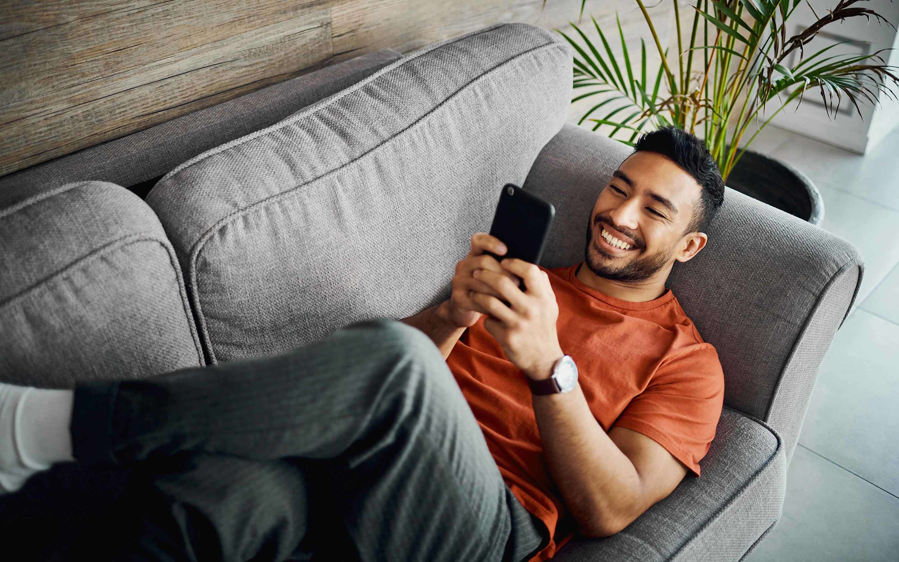 Strom und Gas mit Prämie - So sparst du einfach bei deinem Tarif Junger Mann liegt lächelnd auf dem Sofa und schaut entspannt auf sein Smartphone