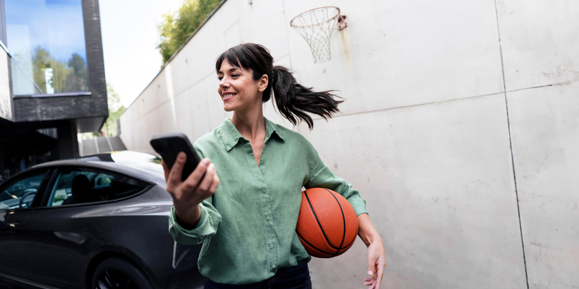 So funktionieren flexible Stromtarife Frau mit Basketball und Handy lädt ihr E-Auto zuhause