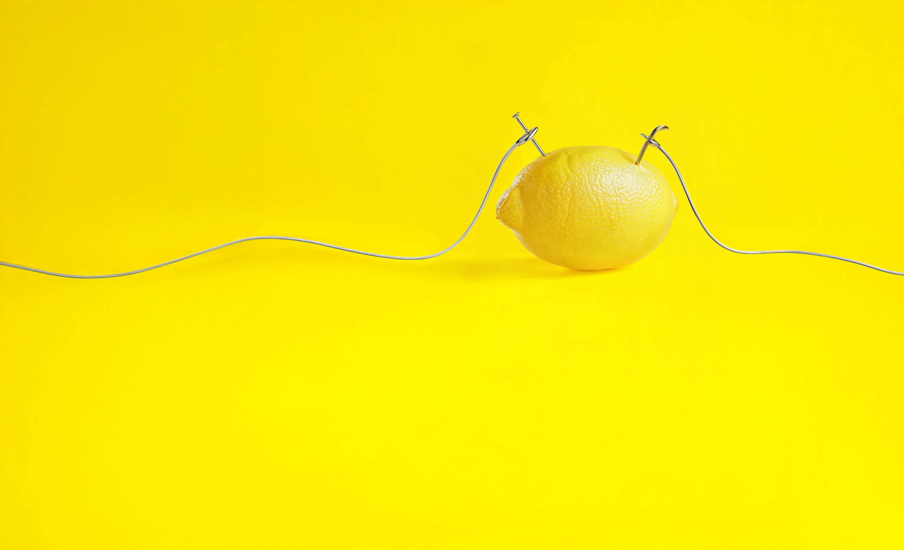 Wir Yellos stehen immer unter Strom - für unsere Kund:innen Gelbe Zitrone mit zwei silbernen Drähten, die seitlich in die Frucht gesteckt sind, vor gelbem Hintergrund