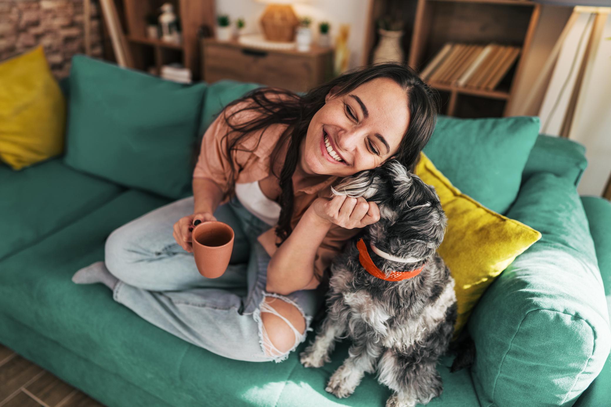 Eine junge Frau lächelt, während sie eine Tasse Kaffee trinkt und mit ihrem Hund auf dem Sofa sitzt