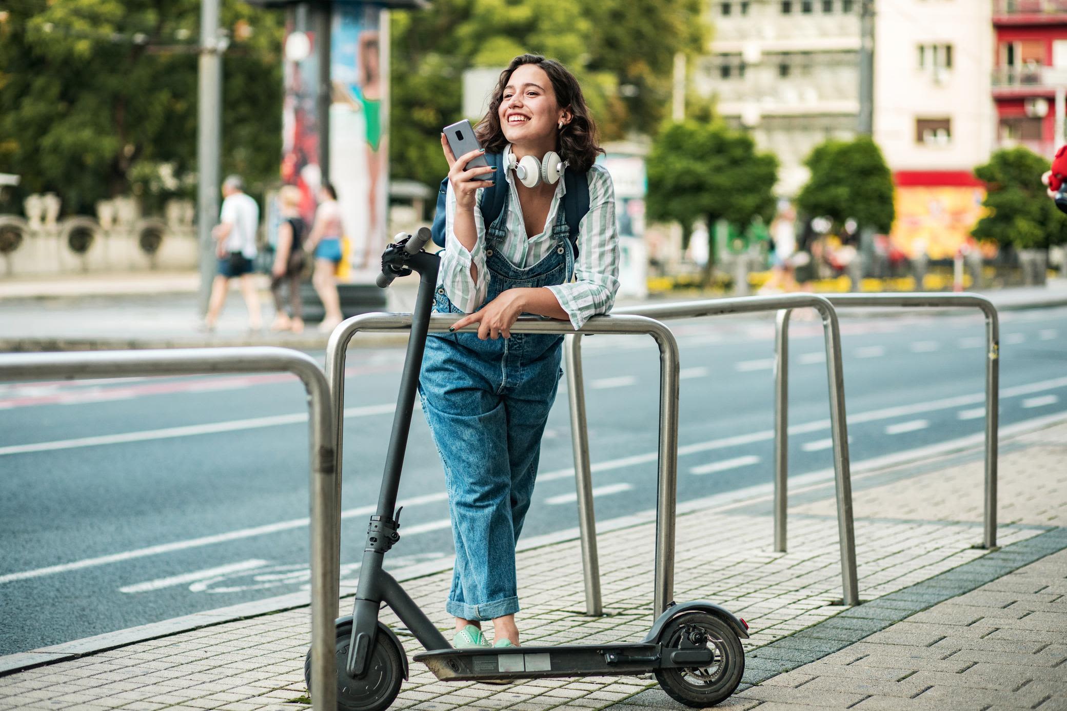 Smart in der Stadt unterwegs mit Elekroscooter Frau mit Smartphone in der Stadt lehnt neben ihrem E-Scooter