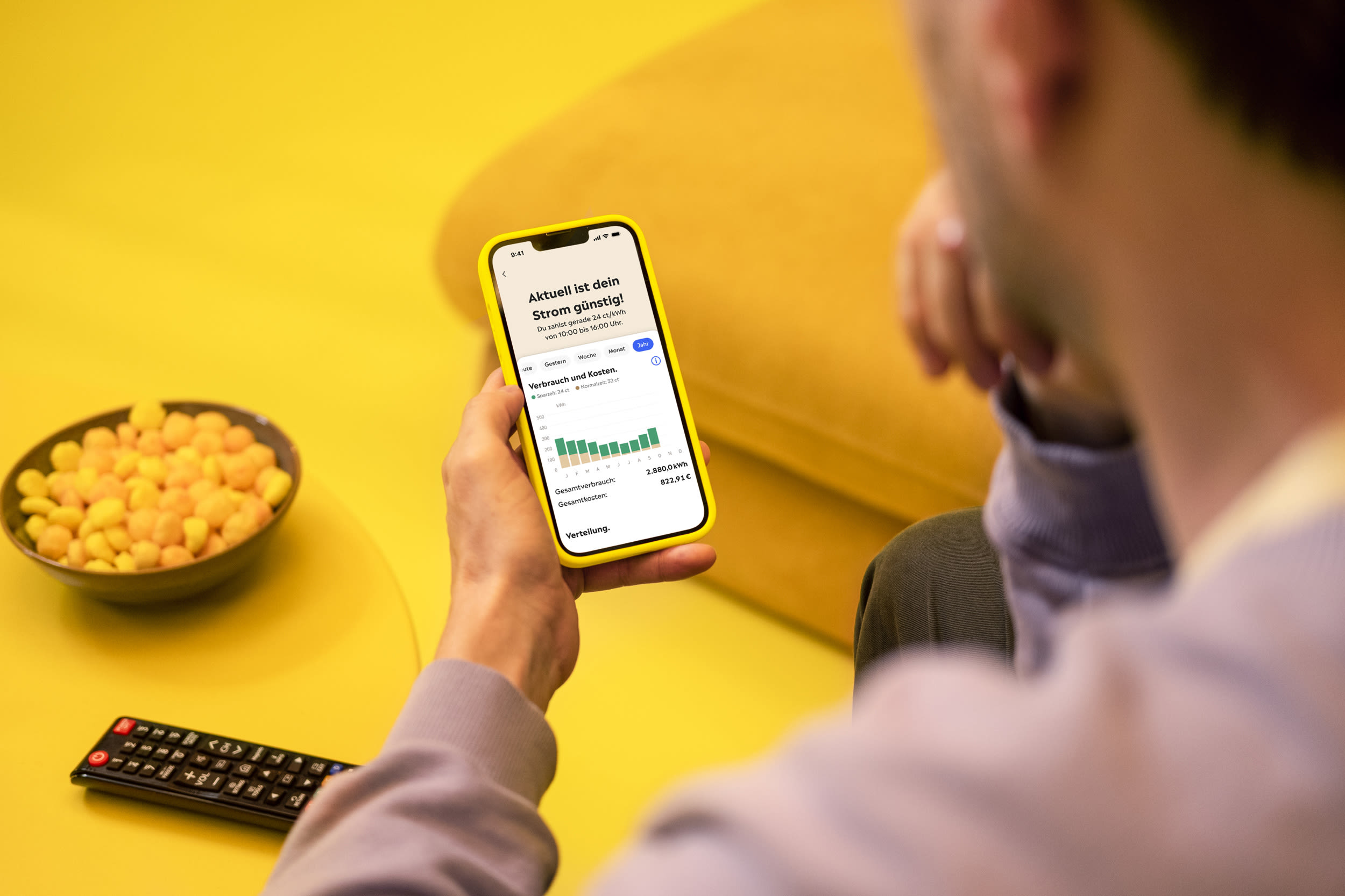 Ein Mann sitzt auf einem gelben Sofa und hält ein Smartphone mit gelbem Rahmen in der Hand. Auf dem Display ist die Yello-App zu sehen, welche die aktuellen Spar- und Normalzeit anzeigt.