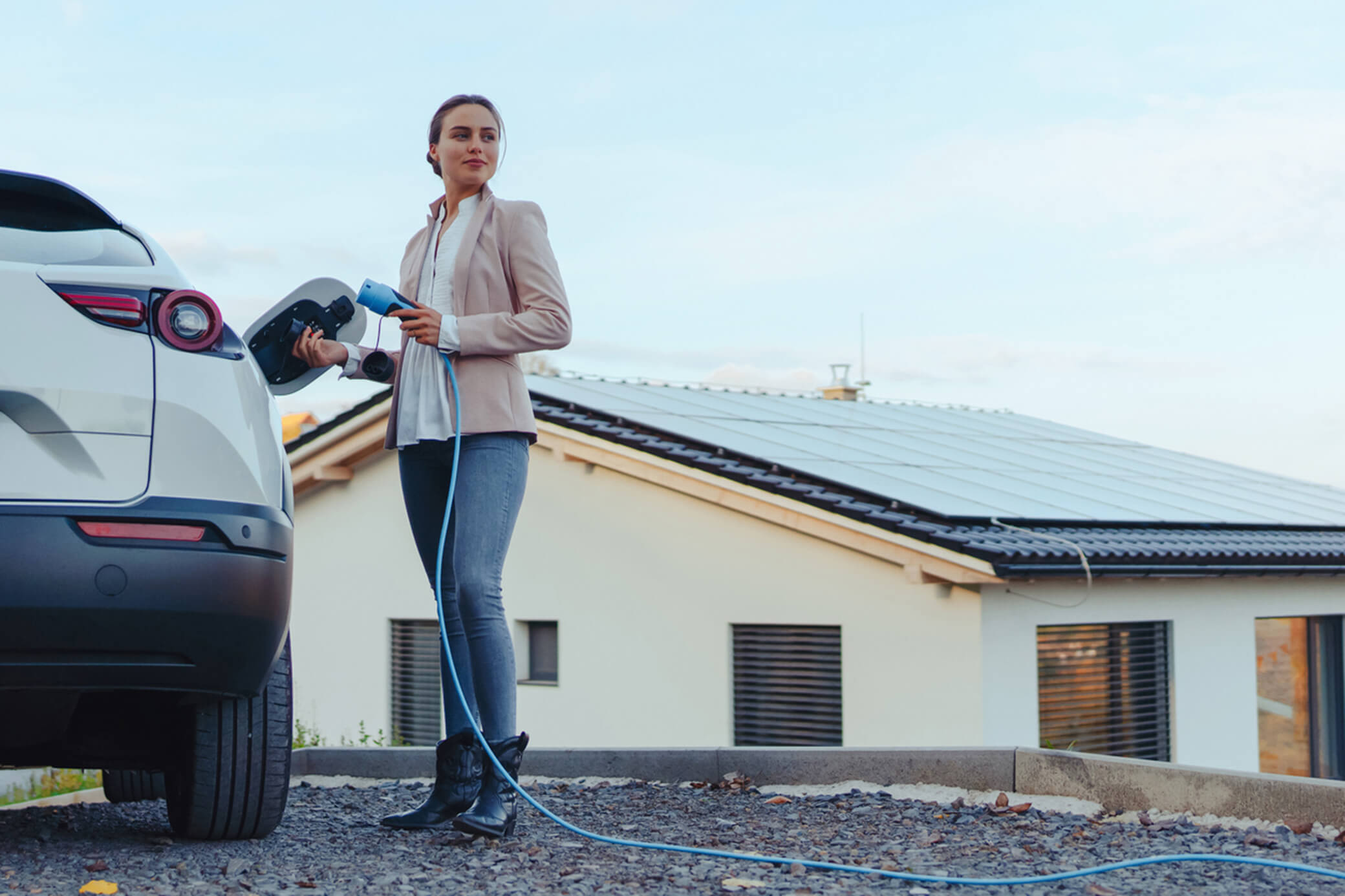 Mit Sonnenstrom lädst du dein E-Auto mit erneuerbarer Energie Eine Frau lädt ein weißes Elektroauto und hält ein Ladekabel in der Hand - im Hintergrund ist eine Solaranlage