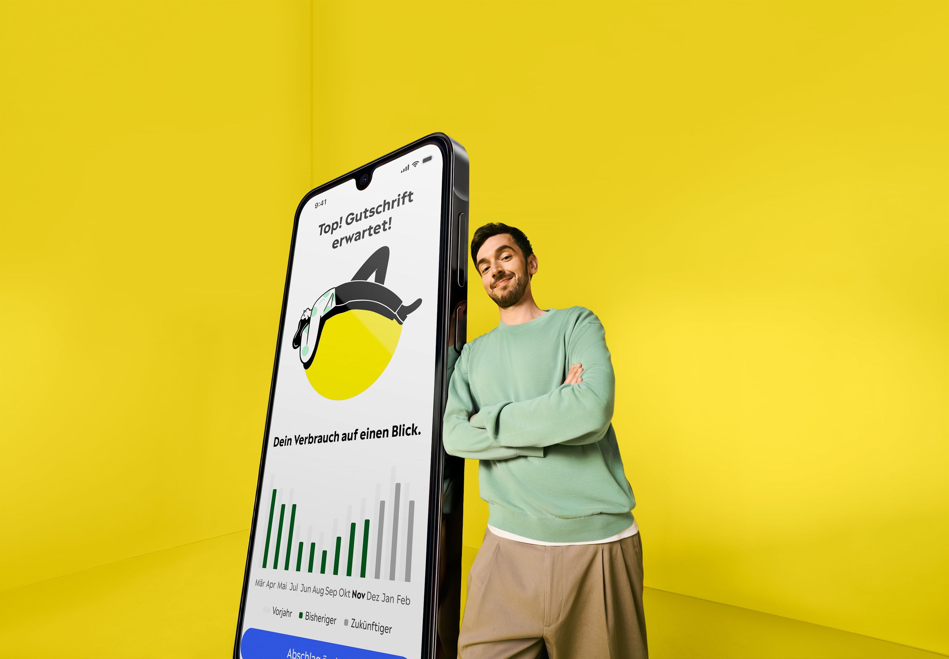 Yello Kampagne Yello Mann steht in einem gelben Raum und lehnt sich an ein übergroßes Smartphone, auf dem die Yello App zu sehen ist.