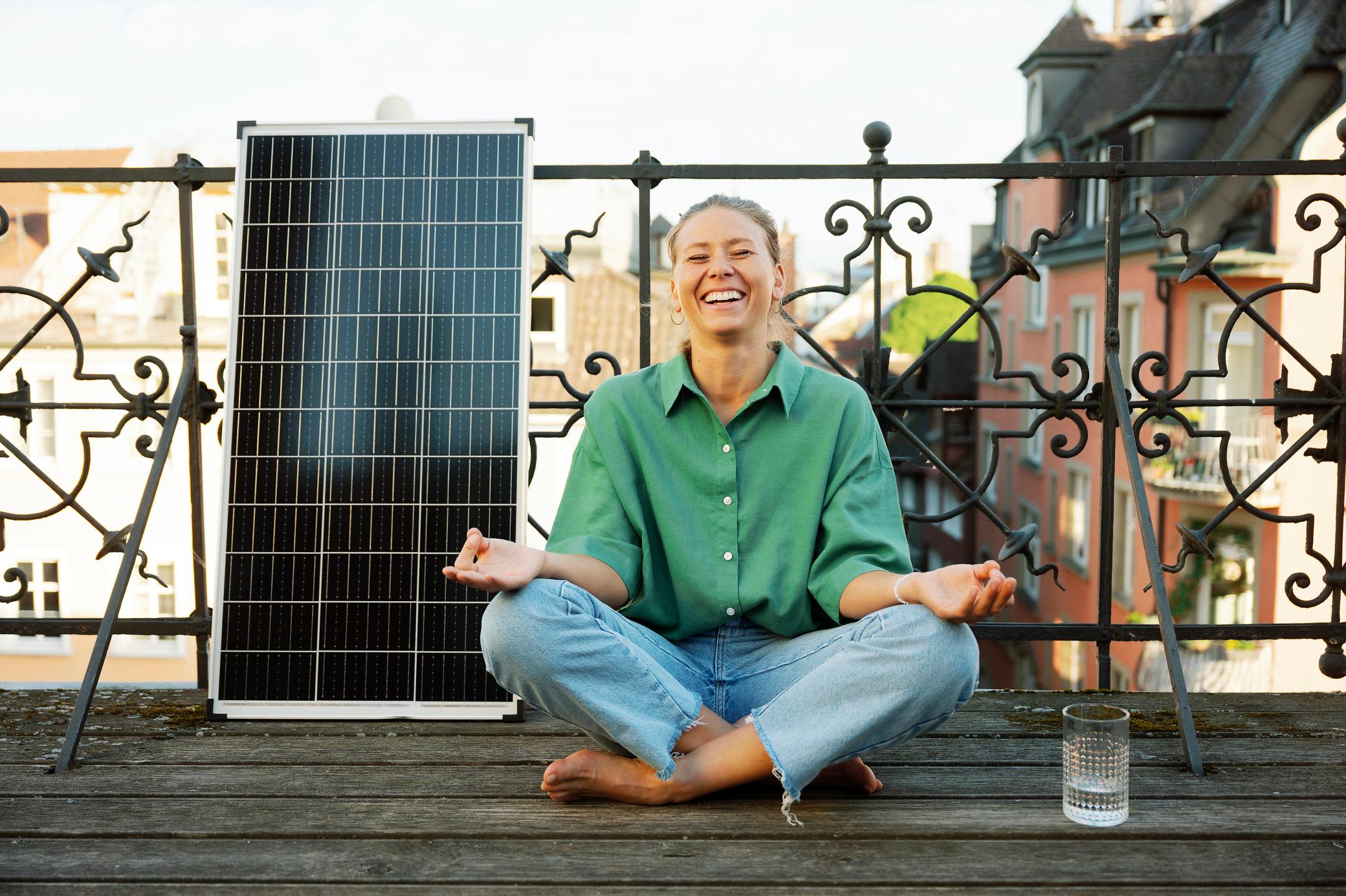 Mit einer Balkon Solaranlage senkst du entspannt Stromkosten. Frau sitzt lachend auf dem Balkon in Yoga-Position, neben einem Balkonkraftwerk
