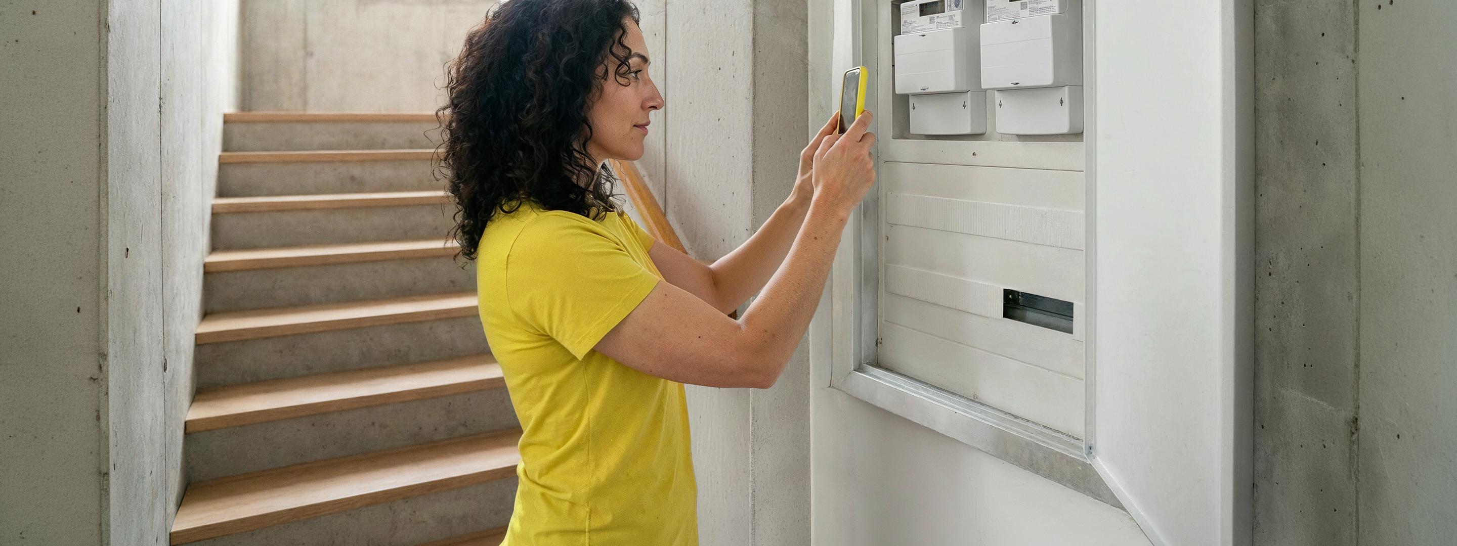 Frau mit braunen Locken und gelbem Shirt steht vor Zählerkasten und fotografiert Zählerstand mit gelbem Handy und der Yello App.