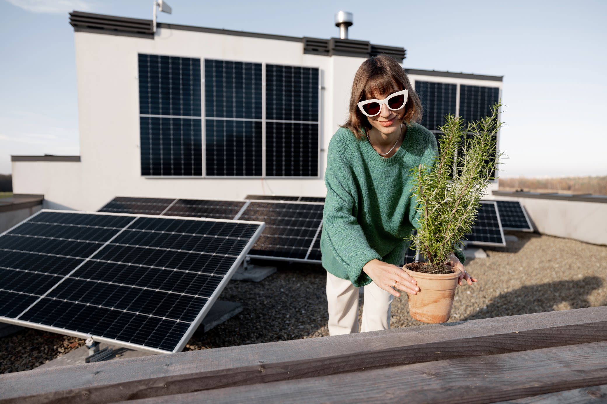 Mit einer eigenen Solaranlage machst du deine eigene Energie Frau mit Sonnenbrille setzt eine Pflanze auf eine Dachterrasse mit Solarmodulen