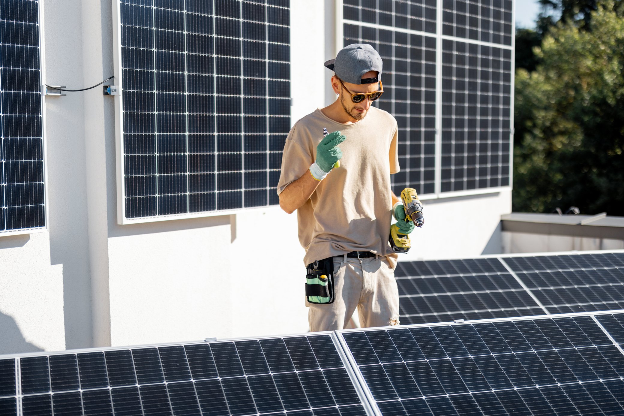 Mann mit Sonnenbrille und Kappe befestigt mit Akkuschrauber Solarmodule auf Balkon.