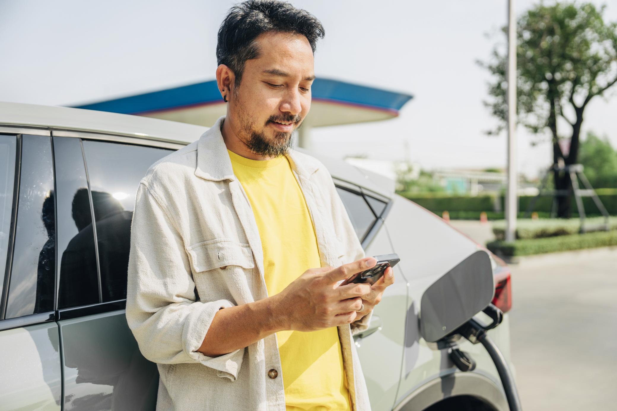 Ein Mann im gelben T-Shirt lädt sein E-Auto unterwegs und schaut aufs Handy