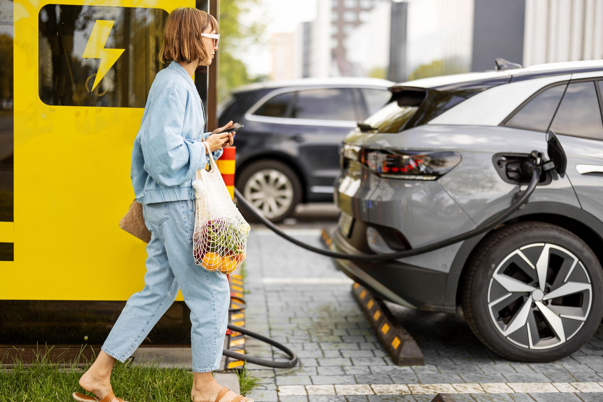 Frau mit Handy und Lebensmitteln wartet auf das Laden ihres Elektroautos in der City