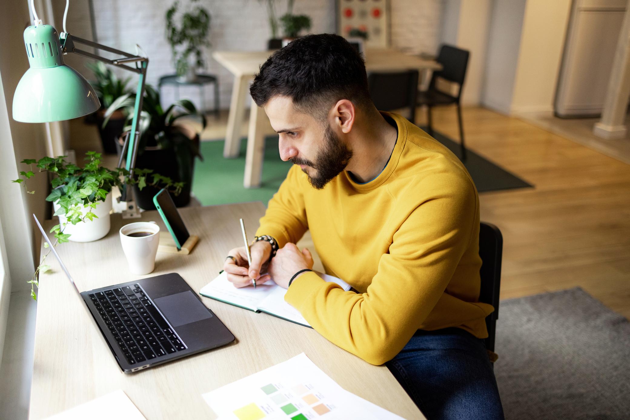 Junger Mann sitzt zuhause am Laptop am Schreibtisch schreibt in sein Notizbuch