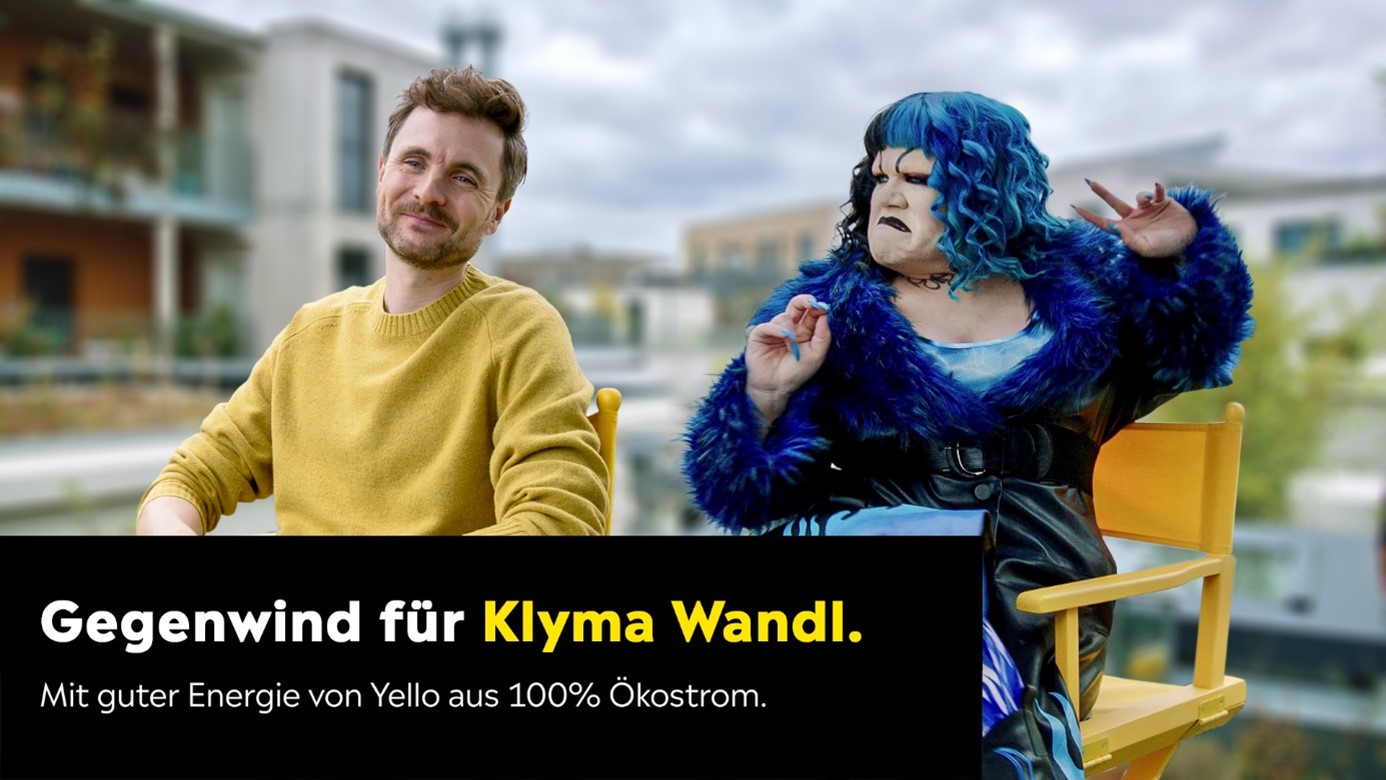 Mann im gelben Pullover und Frau mit blauen Haaren und blauem Mantel sitzen nebeneinander auf Stühlen. Er schmunzelt, während sie grimmig aussieht.
