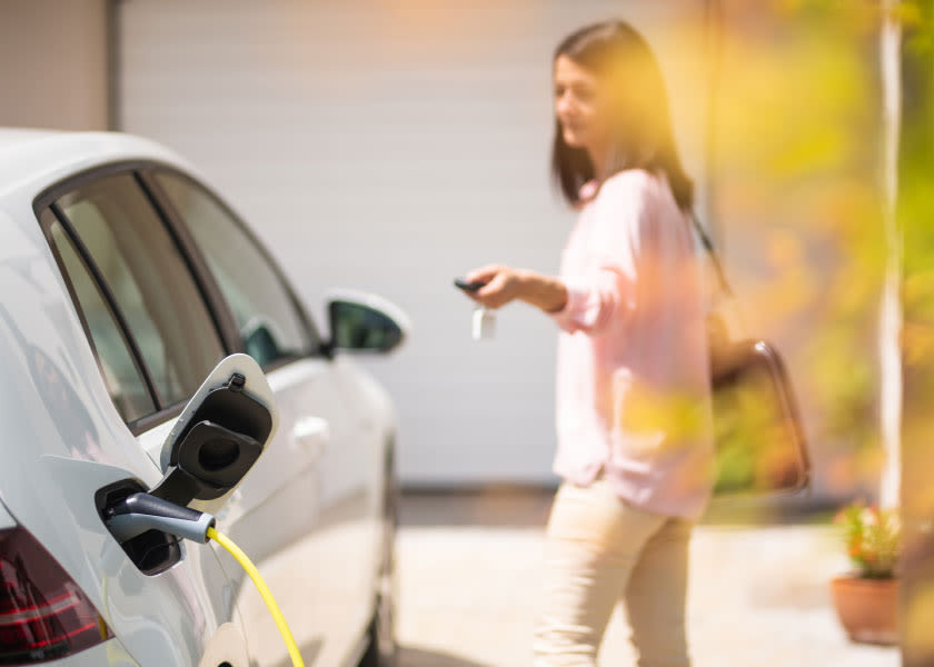 Eine Frau mit Autoschlüsseln steht neben einem weißen Elektroauto, das an ein Ladegerät angeschlossen ist.