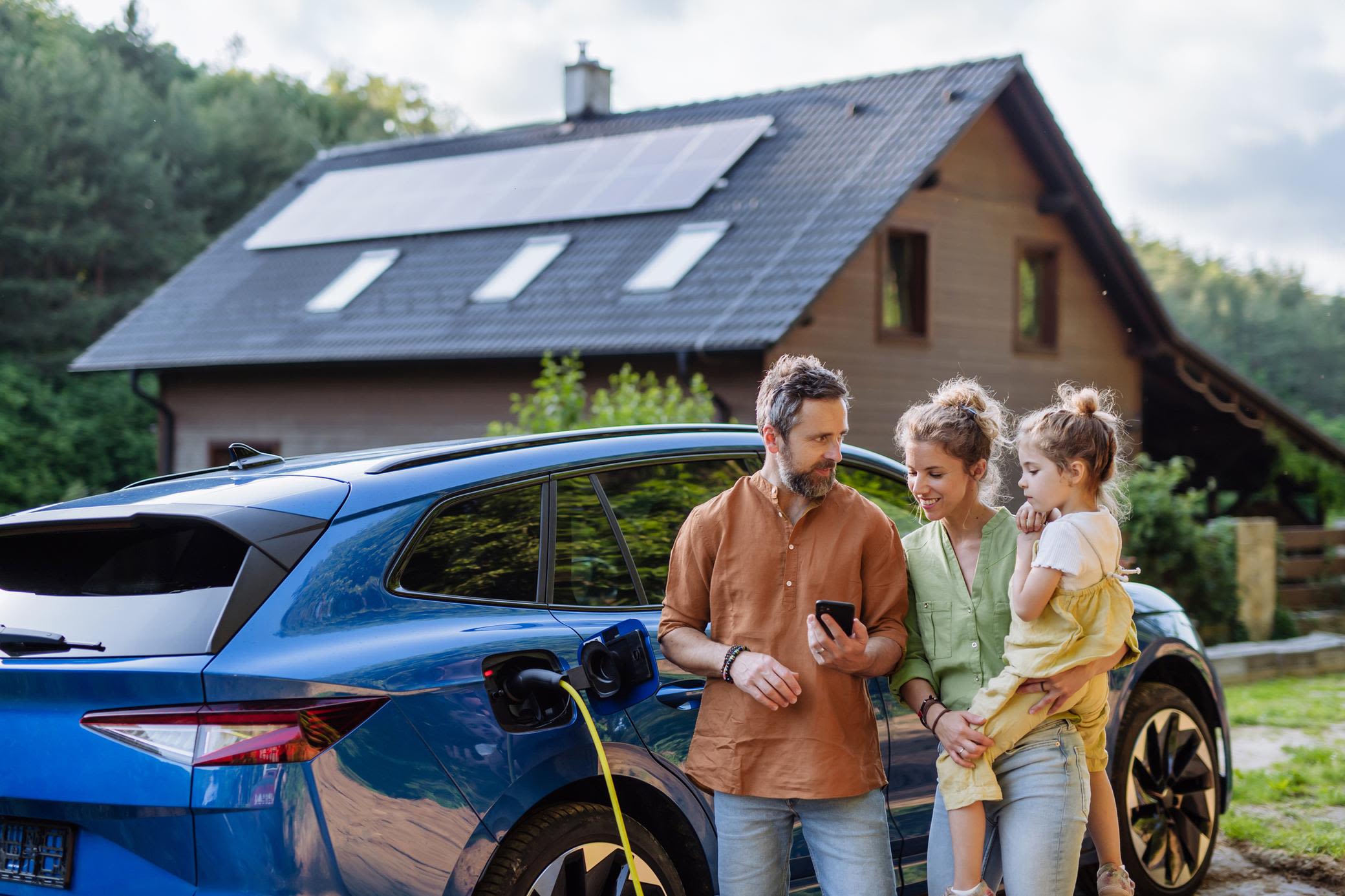 Ob Volleinspeisung oder Eigenbedarf - mit Solar auf dem Dach hat man viele Vorteile und Möglichkeiten Familie mit Mädchen laden Elektroauto vor dem Haus mit Solaranlage auf dem Dach