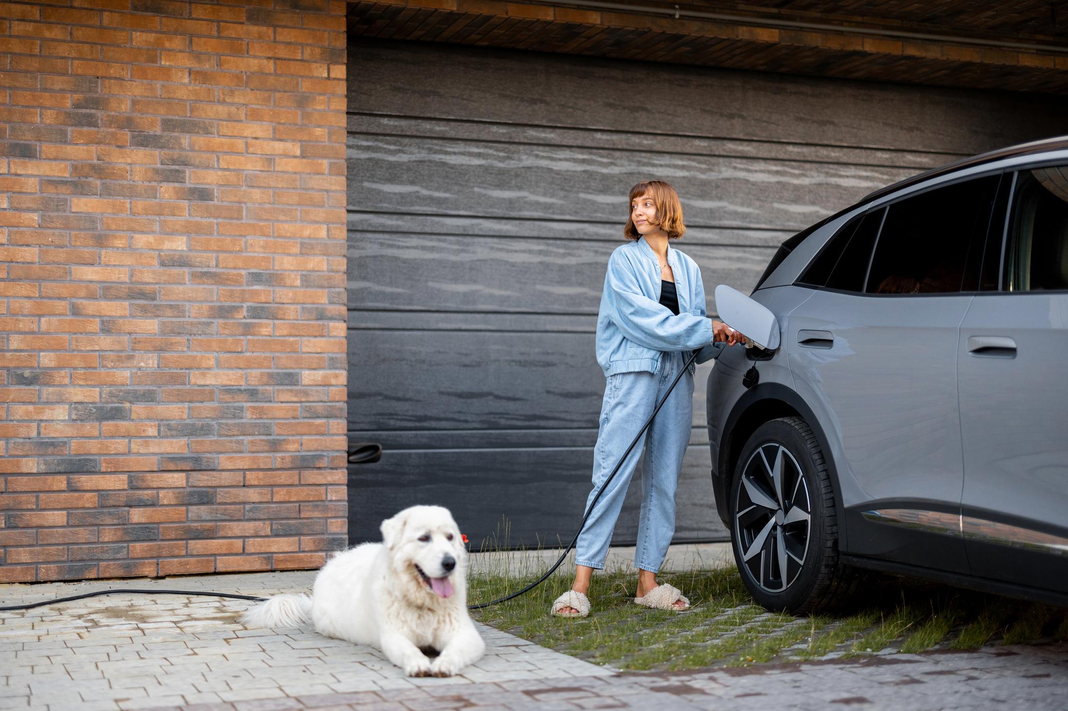 Frau lädt ihr Elektroauto zuhause, ein weißer Hund wartet