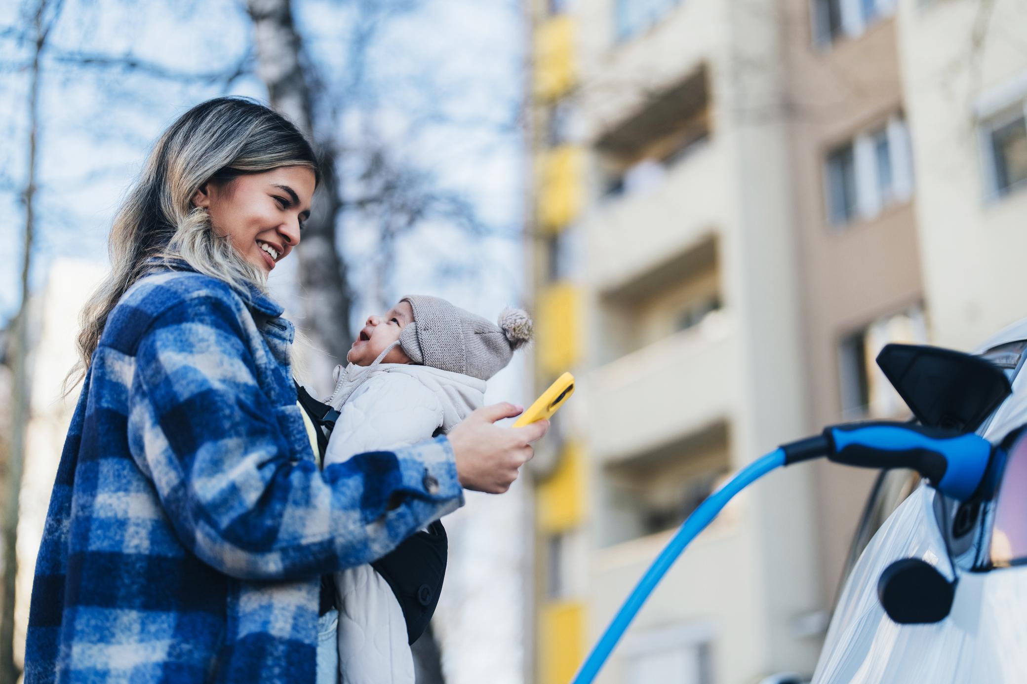 Eine Frau mit Baby in der Trage steht lächelnd an einem E-Auto, das gerade geladen wird und schaut auf ihr Handy. 