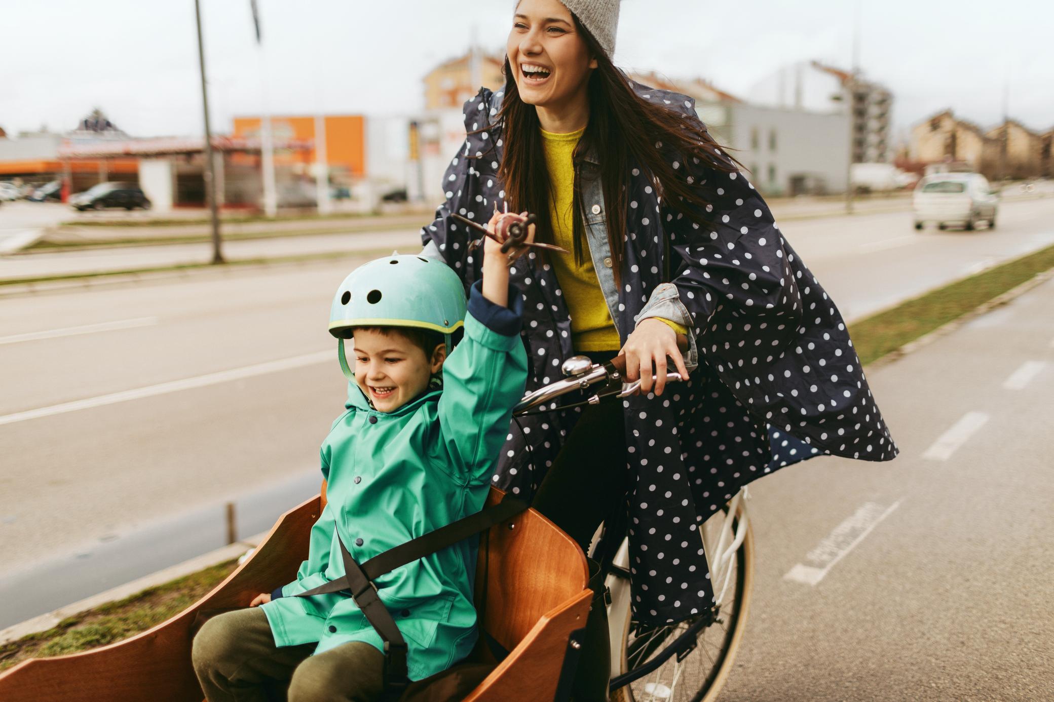 Frau fährt mit ihrem Kind lachend auf dem Fahrrad eine Straße entlang