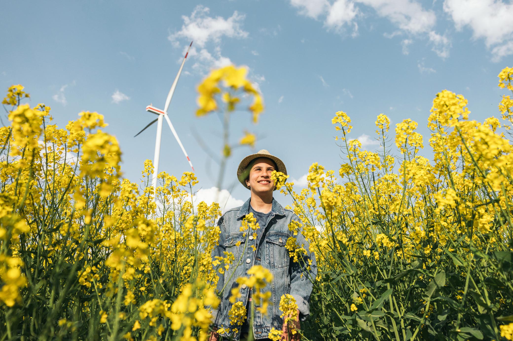 Ein junger Mann steht in einem Feld aus gelben Blumen, hinter ihm ein Windrad