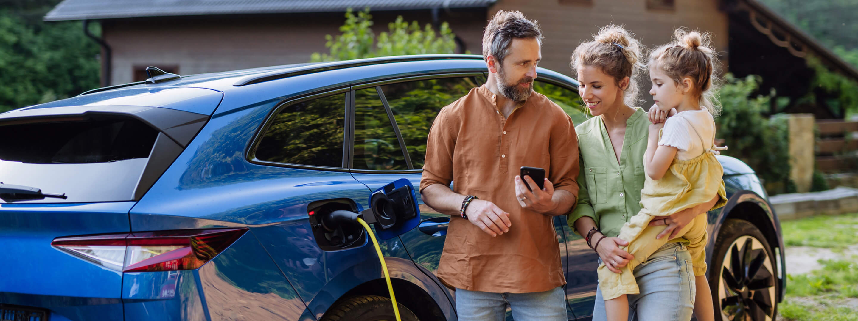 Familie, bestehend aus Vater, Mutter und Tochter, steht vor einem blauen Elektroauto, das gerade geladen wird. Alle blicken auf das Handy des Vaters.