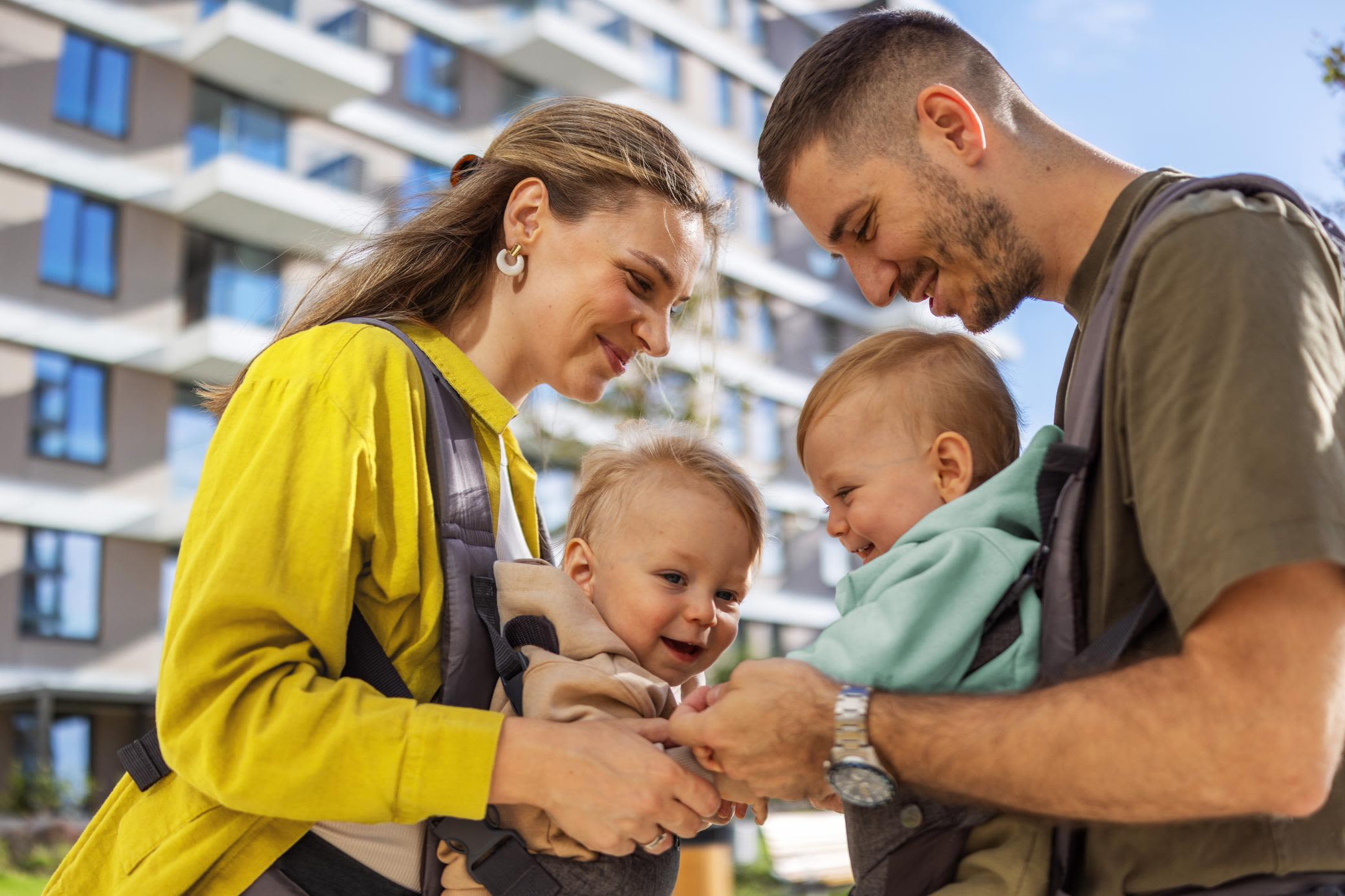 Mit viel guter Energie aus Erneuerbaren geht's in die Zukunft Ein glückliches Paar mit zwei Babys in der Trage steht vor einem Mehrfamilienhaus